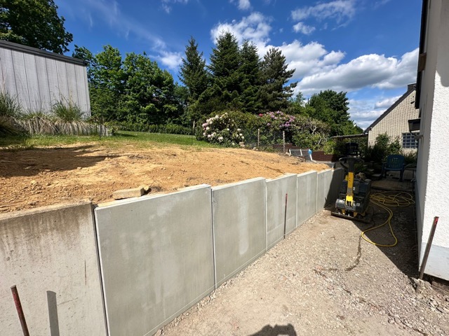 Eine Baustelle mit einer neuen Betonmauer, einem Erdhügel und einem Baugerät unter blauem Himmel.