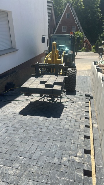 Ein gelber Radlader verlegt Pflastersteine auf einer Baustelle, umgeben von Bäumen und einem Gebäude.