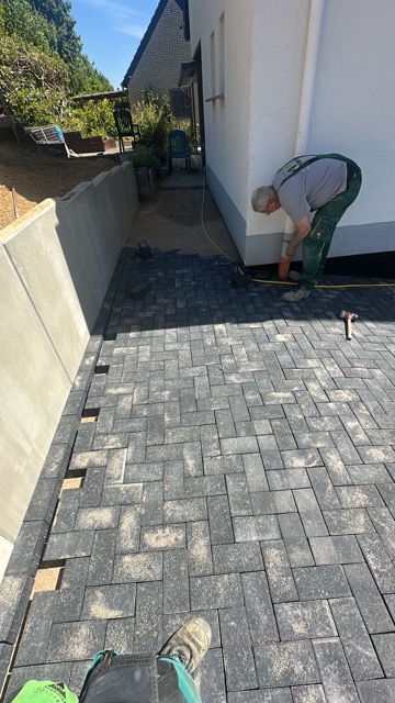 Ein Arbeiter verlegt Pflastersteine auf einer Baustelle, umgeben von neutralen Grautönen und Pflanzen.