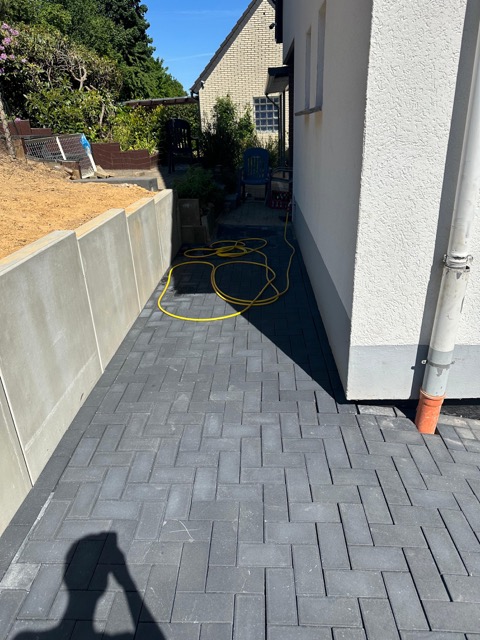 Ein gepflasterter Weg neben einem Haus mit einem gelben Gartenschlauch und einer niedrigen Mauer.