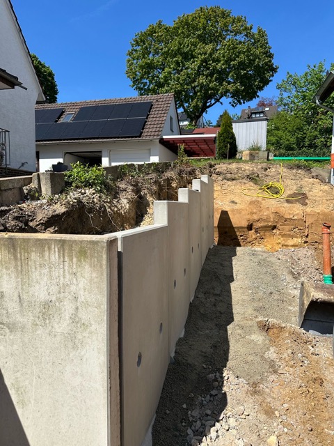 Baustelle in Wohngebiet mit Betonmauer, ausgehobenem Boden, Haus mit Solarpanelen und großem Baum.