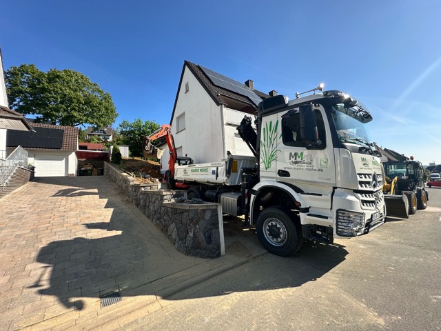 Ein weißer LKW mit Bagger steht in einem sonnigen Wohngebiet, bereit für Bauarbeiten.