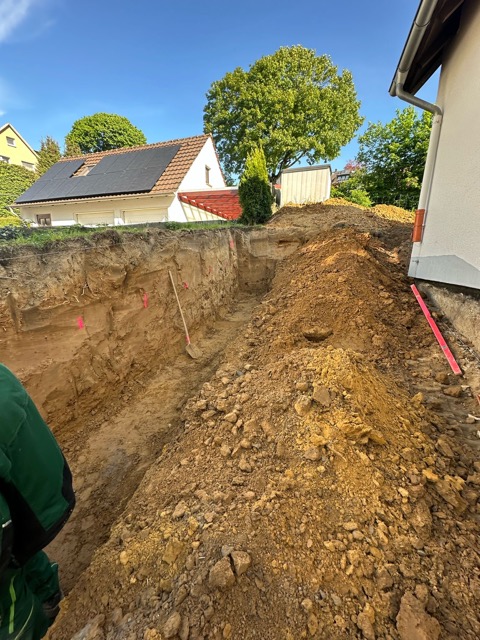 Baustelle in Wohngebiet mit Graben, Erd- und Sandhügeln, Bauarbeiter und Häusern im Hintergrund.