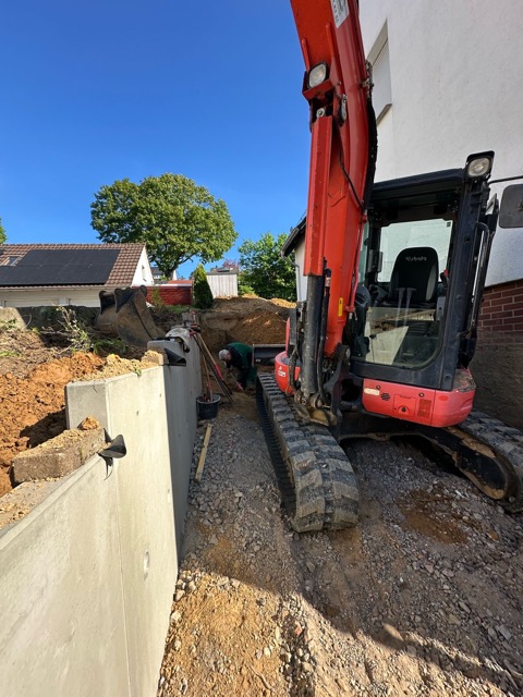 Ein orangefarbener Bagger auf einer Baustelle neben einer Betonmauer, mit einem Haus im Hintergrund.