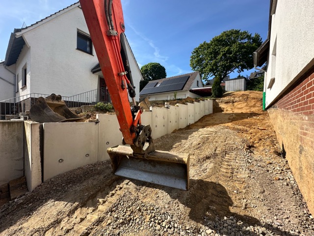 Baustelle mit rotem Bagger und grauen Betonblöcken, umgeben von Wohnhäusern und grünen Bäumen.
