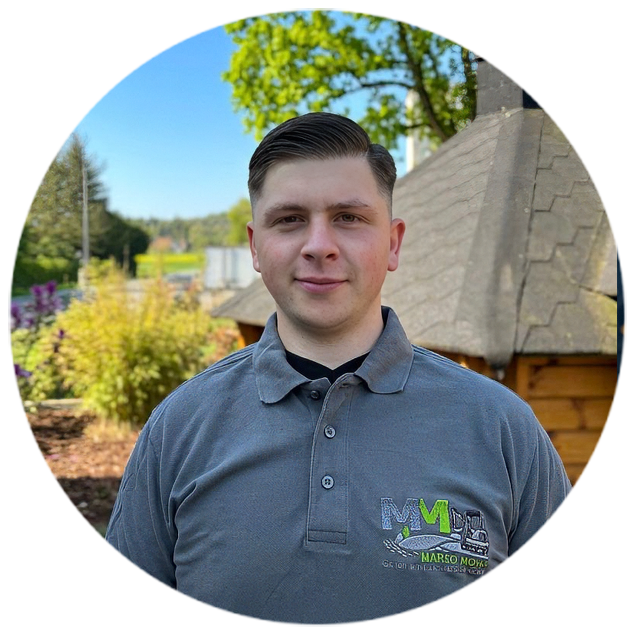 Ein Mann im grauen Polohemd steht vor einem Holzgebäude in einer grünen, ländlichen Umgebung.