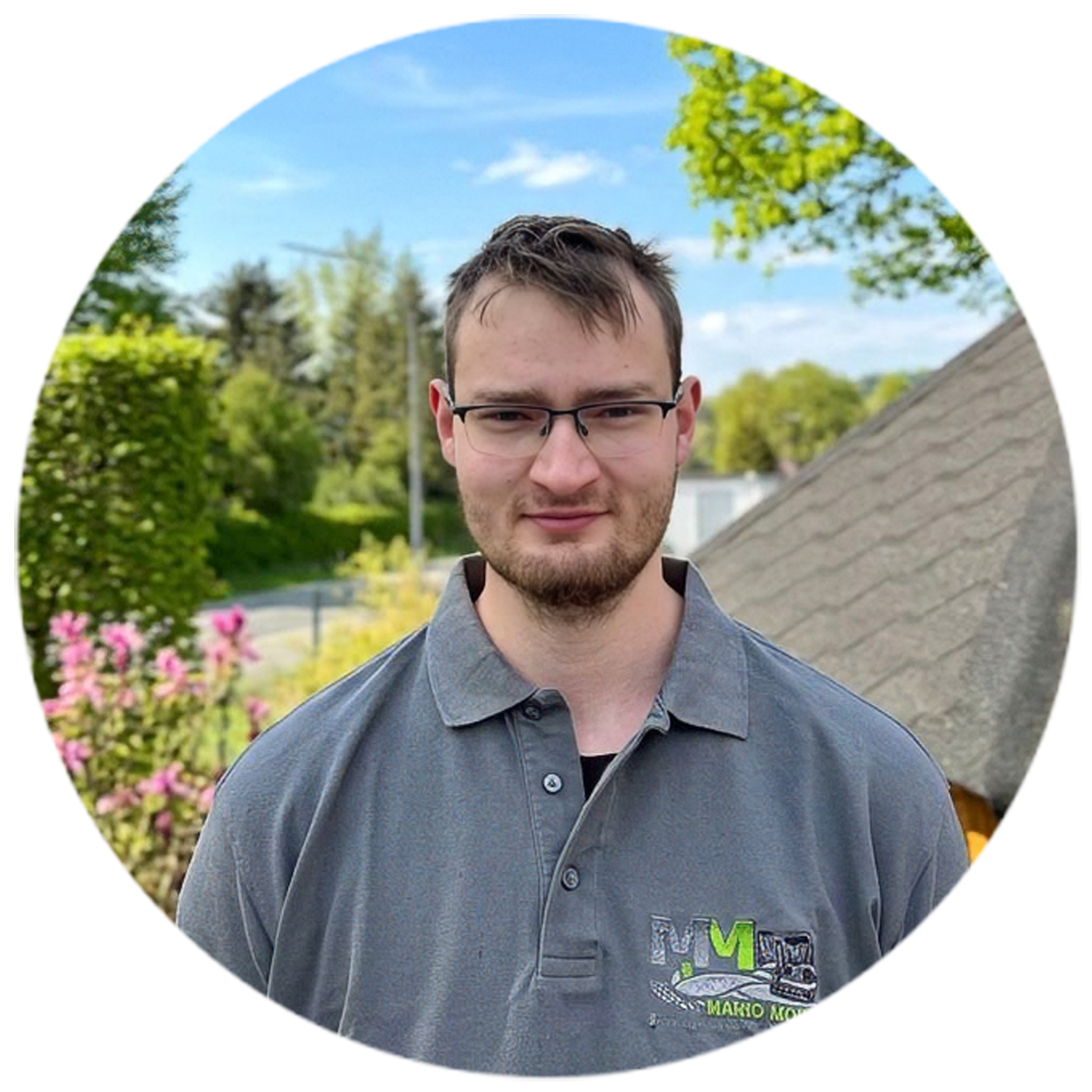 Person im grauen Polohemd mit Brille steht entspannt in grüner, blühender Landschaft unter blauem Himmel.